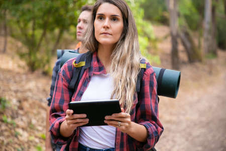 Serious woman checking path via tablet and walking on mountainous trail. Caucasian hikers or...