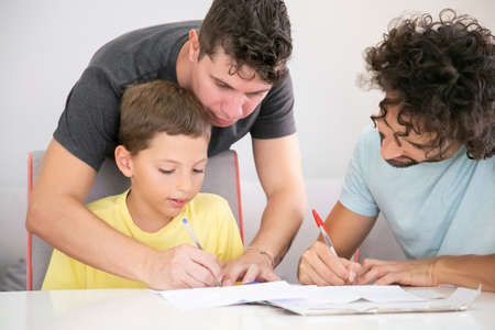 Gay parents helping focused boy with school home task, sitting at table and writing in papers...