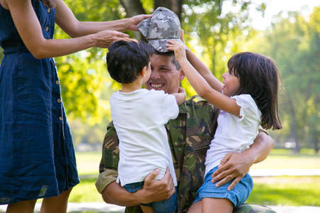 Two happy kids and their mom meeting and hugging military dad in camouflage uniform outdoors. Family...