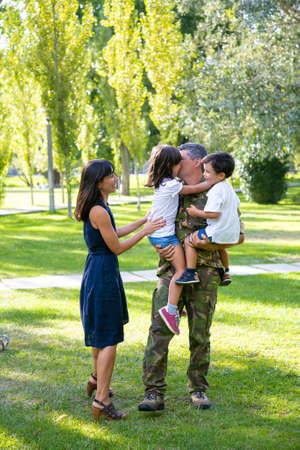 Cheerful mom and two kids meeting military dad in camouflage uniform outdoors. Father taking children in arms and kissing. Family reunion or returning home conceptの写真素材