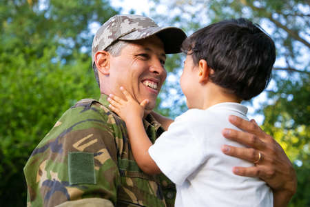 Joyful father holding little son in arms, hugging boy outdoors after returning from military mission...