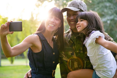 Cheerful military man with wife and little daughter taking selfie on cellphone in city park. Medium...