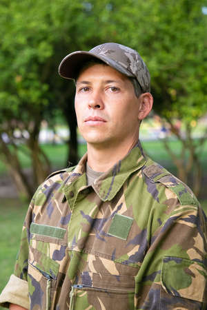 Portrait of serious man in military camouflage uniform standing in park, looking at camera. Green trees in background. Medium shot, low angle. Military man or guard conceptの写真素材