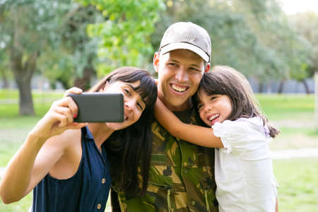 Happy military man, his wife and little daughter taking selfie on cellphone in city park. Front view. Family reunion or returning home conceptの写真素材