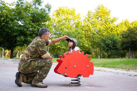 Military dad playing with daughter on playground, dressing girl in camouflage cap while she riding...
