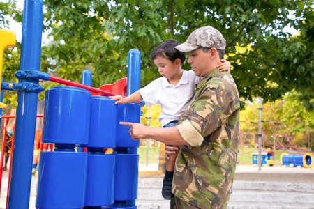 Military dad walking with little son in park, holding boy in arms and studying playground equipment....