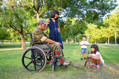 Cheerful mom and disabled military dad in wheelchair looking at kids arranging firewood for campfire outdoors. Disabled veteran or family outdoors conceptの写真素材
