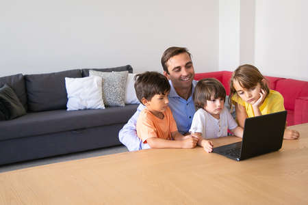 Exited dad and serious kids watching movie via laptop together during quarantine. Smiling father...