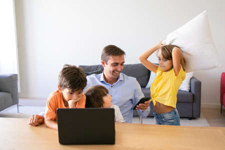 Happy dad using smartphone, sitting at table and kids playing with him. Caucasian father working at...