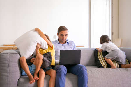 Middle-aged dad sitting on sofa and working on laptop when kids playing near him. Caucasian father...