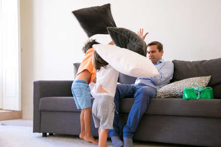 Playful boys attacking Caucasian man with pillows. Cheerful children playing and having fun with his father. Dad closing eyes and defending from attack. Childhood, family and game activity conceptの写真素材