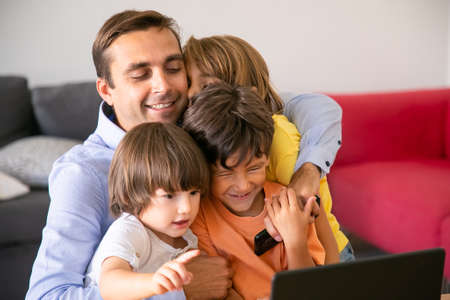 Happy father hugging with cute children. Caucasian middle-aged dad sitting in living room, embracing cute kids, holding mobile phone and smiling. Fatherhood, childhood and family conceptの写真素材