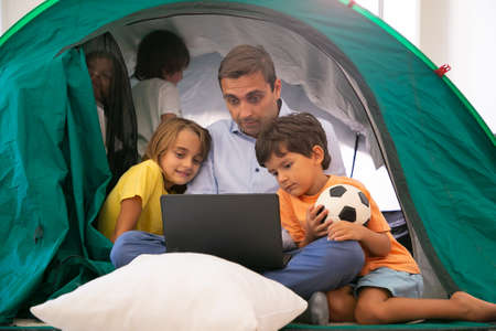 Caucasian dad sitting cross-legged with kids in tent at home and watching movie via laptop. Lovely...
