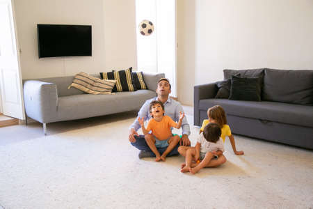 Exited father sitting on carpet with children and playing. Cute playful boy throwing up ball and...