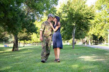 Loving Caucasian couple embracing, kissing and walking together on lawn in park. Middle-aged soldier in military uniform, hugging his pretty wife. Family reunion, weekend and returning home conceptの写真素材