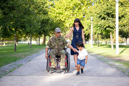 Serious military father walking in wheelchair with family. Caucasian middle-aged dad in camouflage...