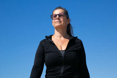 Focused confident mature woman in sportswear walking outside. Low angle, clear blue sky in...