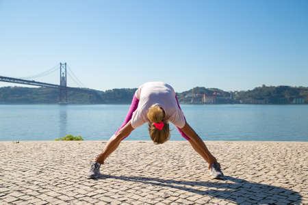 Mature woman in sports clothes doing morning exercise outside, stretching back and legs muscles at...
