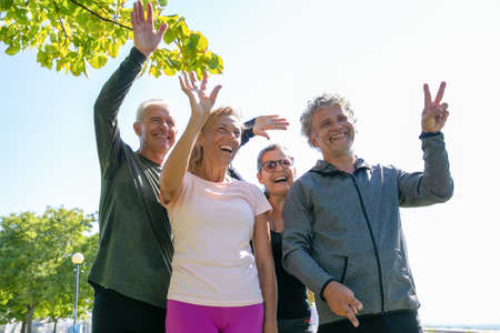 Happy cheerful mature people in sports clothes standing together in park, looking away and waving...