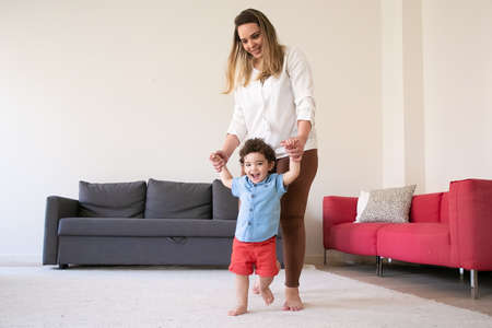Happy mother holding son hands and teaching him to walk. Cheerful curly mixed-race little boy walking on carpet barefoot with help of long-haired mom. Family time, childhood and first step conceptの写真素材
