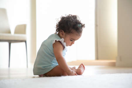 Pensive dark curly haired baby girl wearing pale blue dress, sitting on floor at home, touching carpet, looking down. Side view. Kid at home and childhood conceptの写真素材
