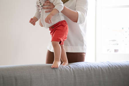 Mother helping baby to walk on couch at home. Kid making first steps with moms support. Cropped...