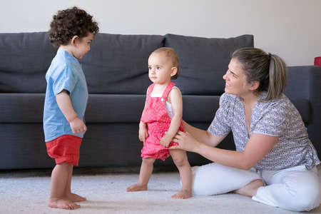 Happy mother playing with little children at home and sitting cross-legged. Cute baby girl and boy standing barefoot on carpet in living room. Family indoors, weekend and childhood conceptの写真素材