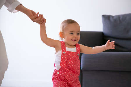 Cheerful funny infant learning walking in living room and holding mother hand. Cute baby girl playing with unrecognizable mother at home and smiling. Childhood, weekend and first step conceptの写真素材