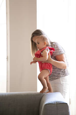 Mom helping baby to walk at home. Kid making first steps with moms support. Side view. Parenthood and childhood conceptの写真素材