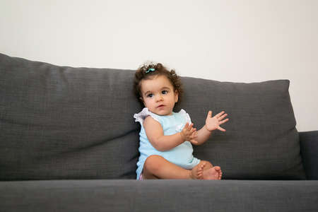 Adorable dark curly haired baby girl in pale blue cloth sitting on grey couch at home, looking away and clapping hands. Kid at home and childhood conceptの写真素材