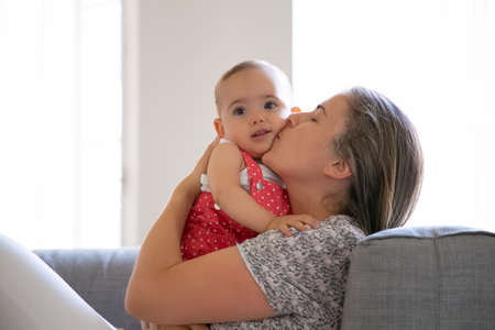 Caring mother sitting on sofa and kissing her little daughter with love. Adorable baby girl looking at camera. Long-haired Caucasian mum holding infant with both hands. Family and motherhood conceptの写真素材