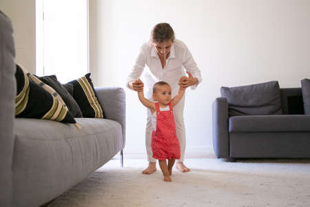Front view of mom holding little girl hands and helping to walk. Cute barefoot baby girl in red...