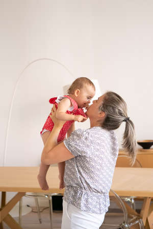 Blonde mother playing with cute baby girl, rising her up and having fun. Happy infant in red dungarees shorts looking at mom and biting finger. Family time, motherhood and being at home conceptの写真素材