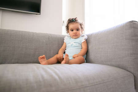 Serious dark curly haired baby girl wearing pale blue cloth, sitting on grey couch at home, looking at camera. Kid at home and childhood conceptの写真素材