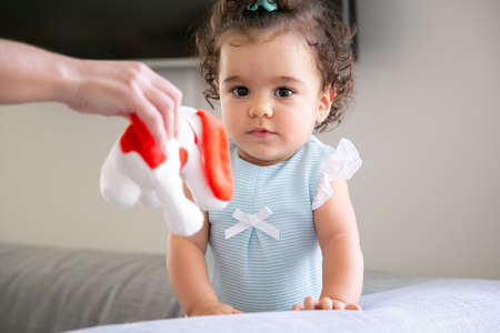Pensive dark curly haired baby girl looking at camera while her mom giving red and white toy to child. Kid at home and childhood conceptの写真素材