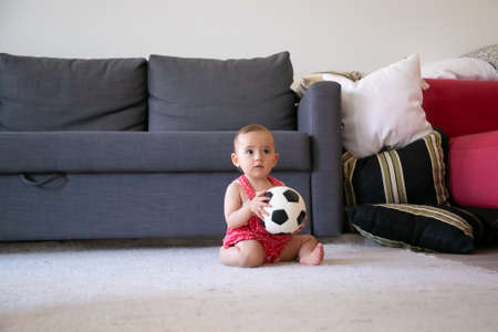 Thoughtful baby girl holding soccer ball, sitting on carpet barefoot and looking away. Lovely toddler in red dungarees shorts playing at home near sofa. Holiday, weekend and childhood conceptの写真素材