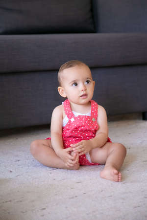 Lovely little baby sitting on carpet barefoot in living room. Cute thoughtful girl in red dungarees shorts looking at someone and touching foot. Weekend, childhood and being at home conceptの写真素材