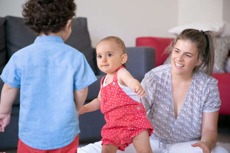 Happy kids playing in living room with blonde mom. Cute little girl standing and looking at curly boy. Smiling loving mother watching children. Family indoors, weekend and motherhood conceptの写真素材