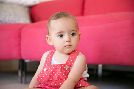 Pensive baby sitting on floor in living room and thinking about something. Serious little girl in red dungarees shorts looking away and sitting near sofa. Weekend, childhood and being at home conceptの写真素材