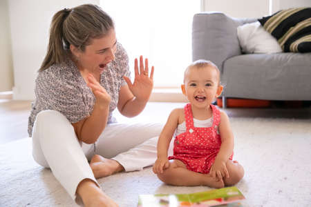 Exited little girl sitting on floor and playing with mother. Cheerful blonde mom having fun with her...