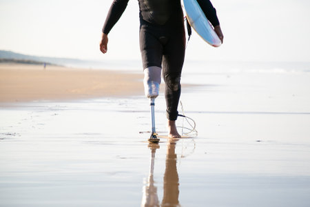 Unrecognizable surfer with leg prosthesis walking near sea. Strong man wearing black swimsuit, carrying surfboard and going along coastline. Artificial limb, lifestyle and extreme sport conceptの写真素材