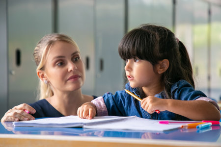 Serious young female teacher helping primary school girl to do her task. Little girl pointing at her open copybook and speaking. Education or back to school conceptの写真素材