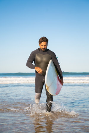 Pensive handicapped surfer walking in sea water with board. Bearded middle-aged amputee with artificial leg carrying surfboard and looking down. Physical disability and extreme sport conceptの写真素材