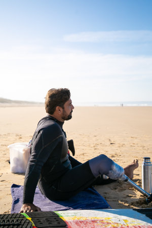Middle-aged surfer with artificial leg sitting on beach and looking at sea. Attractive bearded amputee relaxing alone after surfing. Physical disability, lifestyle and extreme sport conceptの写真素材