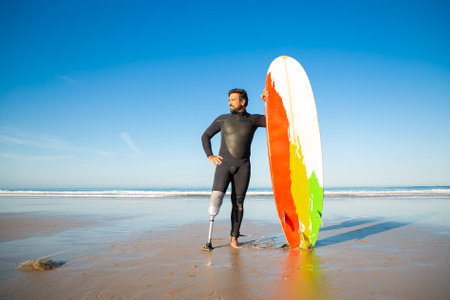 Confident handicapped man standing on sea beach with board. Attractive brunette man with artificial leg wearing black wetsuit and looking at ocean. Physical disability and extreme sport conceptの写真素材