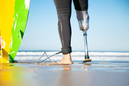 Closeup view of artificial leg of disabled surfer standing near surfboard. Feet of male amputee on sea coast. Ocean waves on background. Physical disability, lifestyle and extreme sport conceptの写真素材