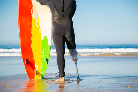 Back view of unrecognizable amputee standing with surfboard on beach. Handicapped man with amputated leg looking at ocean and wearing swimsuit. Physical disability, lifestyle and extreme sport conceptの写真素材