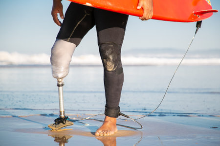 Cropped male surfer standing with surfboard on sea beach. Unrecognizable amputee with artificial leg walking along coastline with board. Physical disability, lifestyle and extreme sport conceptの写真素材