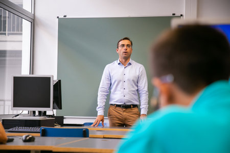 Serious male teacher conducting class, standing at blackboard, explaining task to students....