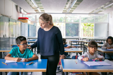 Serious school teacher watching pupils doing their task in class, sitting at desks and writing in copybooks. Education or back to school conceptの写真素材